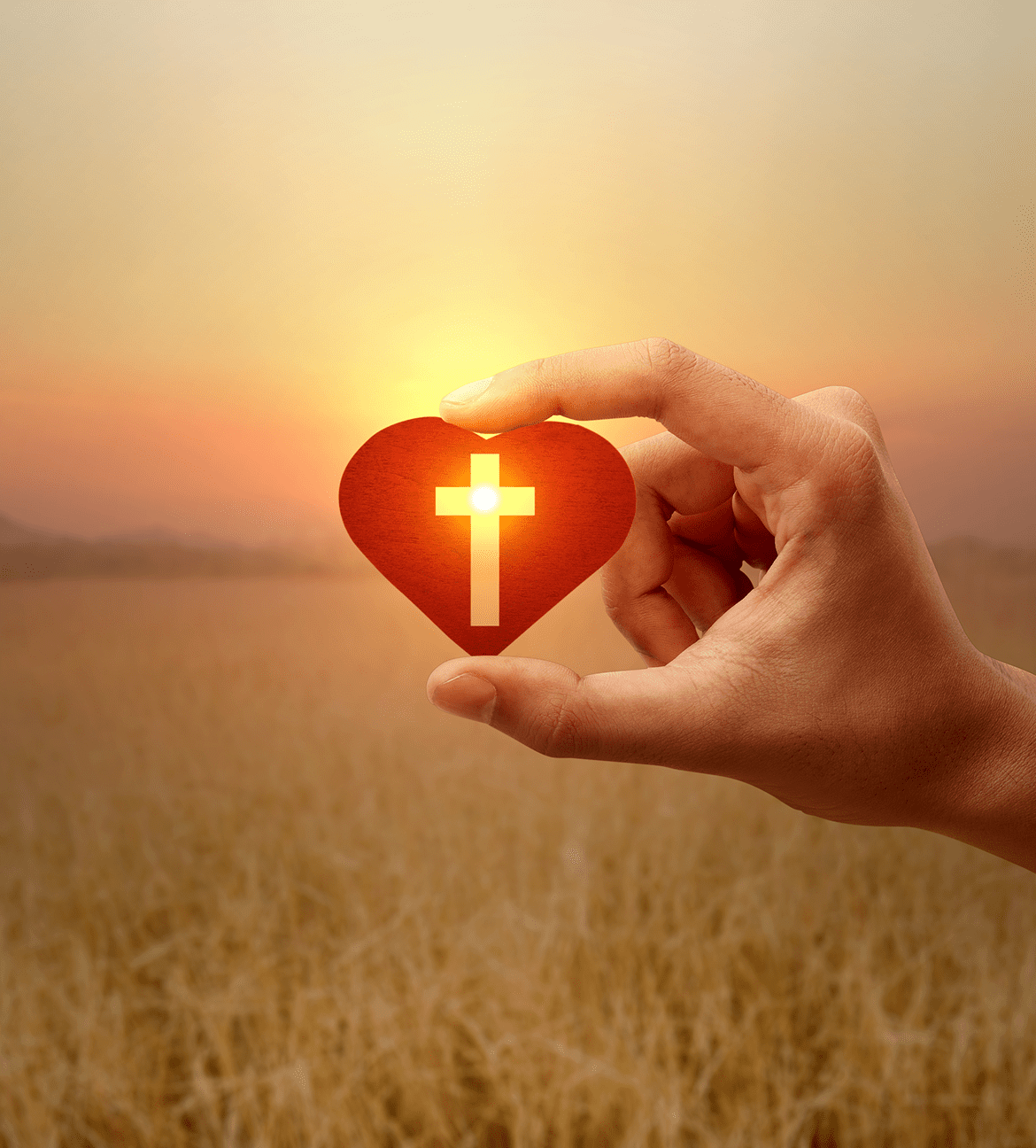 Hand holding heart with cross at sunset.